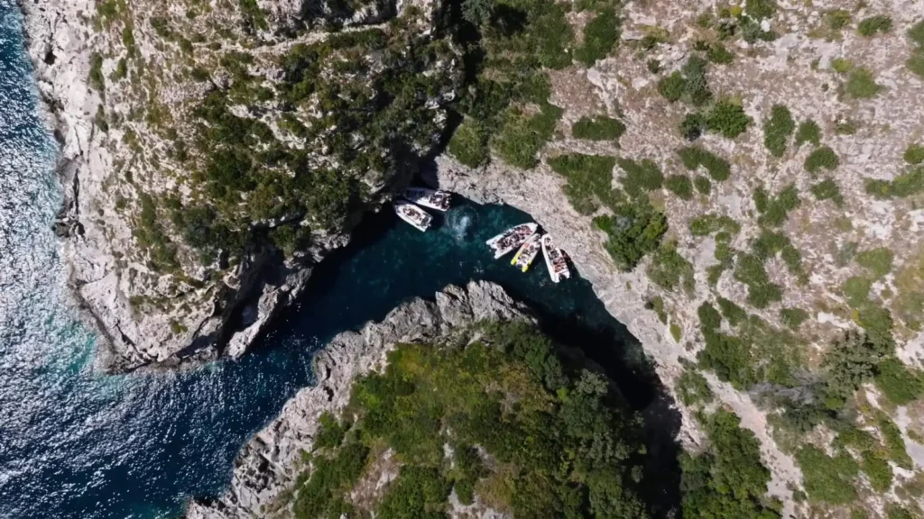 Aerial view of Englishman Bay on the Karaburun Peninsula, showing small boats anchored in a narrow rocky inlet surrounded by rugged limestone cliffs and patches of green vegetation above deep blue water.