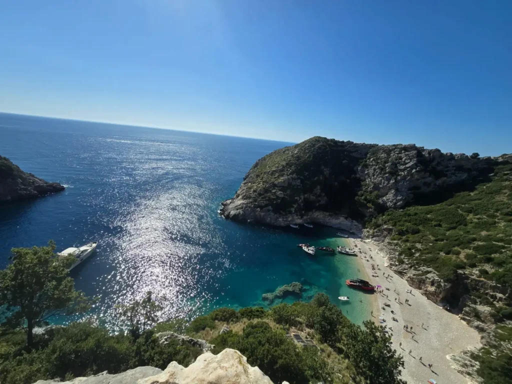 Panoramic view of Grama Bay on the Karaburun Peninsula, featuring a curved pebble beach with visitors and boats anchored in crystal-clear turquoise water, surrounded by rocky cliffs and the open Ionian Sea under a clear blue sky.