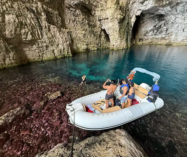 Visitors inside Haxhi Ali Cave in Vlore, standing near a white speedboat anchored in crystal-clear blue water, surrounded by dramatic limestone walls and the rocky interior of the large sea cave.
