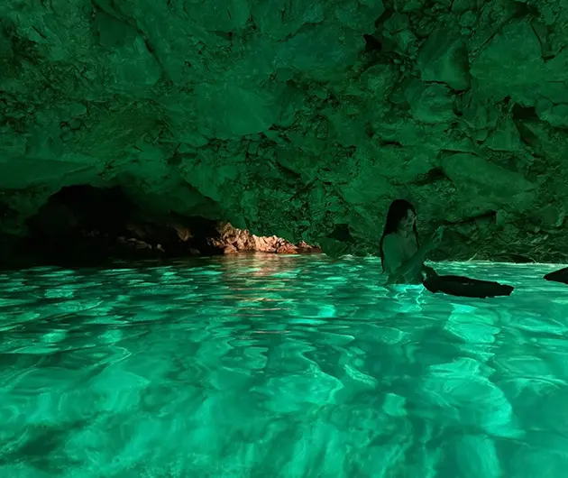 Interior view of the Blue Cave near Vlore, with emerald-green water reflecting sunlight onto the rocky ceiling and a visitor swimming in the illuminated crystal-clear sea inside the cave.