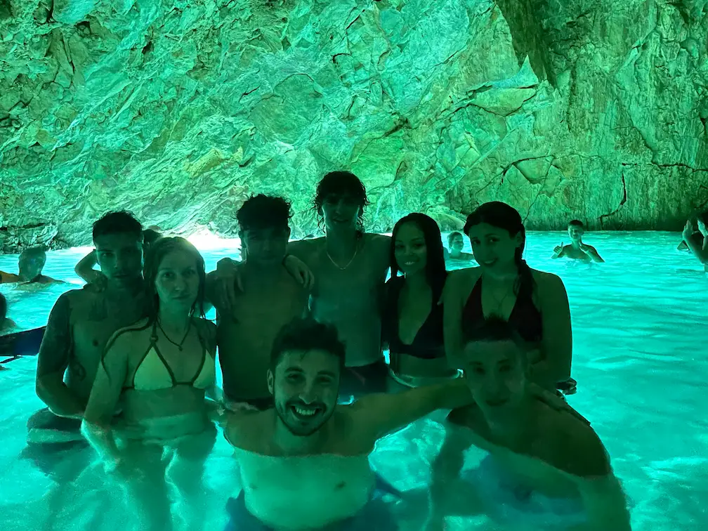 Group of visitors standing together in the glowing turquoise water inside the Blue Cave near Vlore, with dramatic limestone walls illuminated by natural light reflecting across the crystal-clear sea.