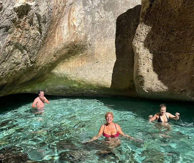 Visitors swimming in the Canyon of Smugglers on the Karaburun Peninsula, with clear turquoise water reflecting sunlight against smooth limestone canyon walls and shaded rock formations.