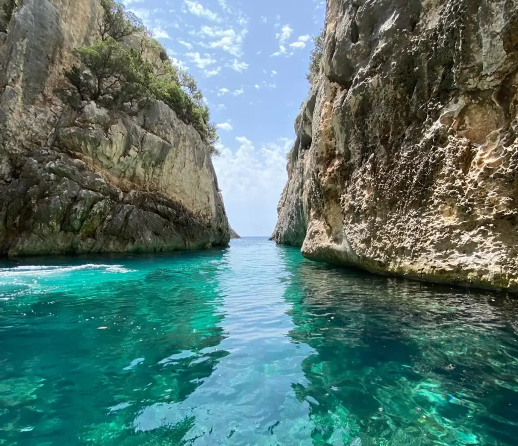Narrow sea passage at the Canyon of Smugglers on the Karaburun Peninsula, with towering limestone cliffs on both sides and crystal-clear emerald water leading toward the open Ionian Sea under a bright blue sky.