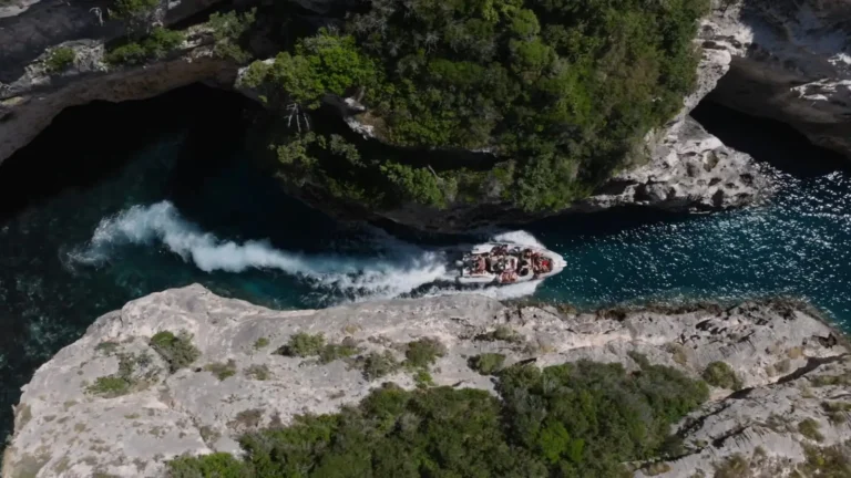 Aerial view of a speedboat navigating through the narrow Canyon of Smugglers on the Karaburun Peninsula, leaving a white wake between steep rocky cliffs and dark emerald water below.