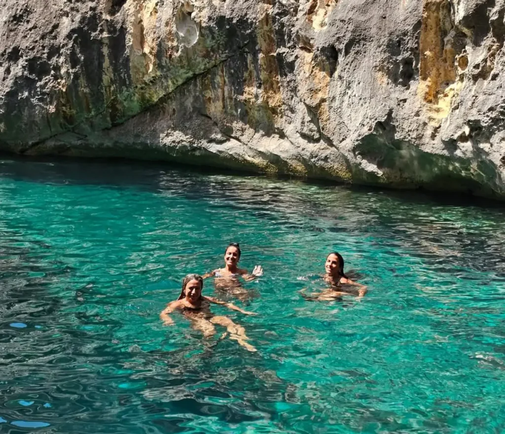 Three visitors swimming in the clear emerald water of the Canyon of Smugglers on the Karaburun Peninsula, with textured limestone cliffs rising steeply behind them along the narrow sea passage.