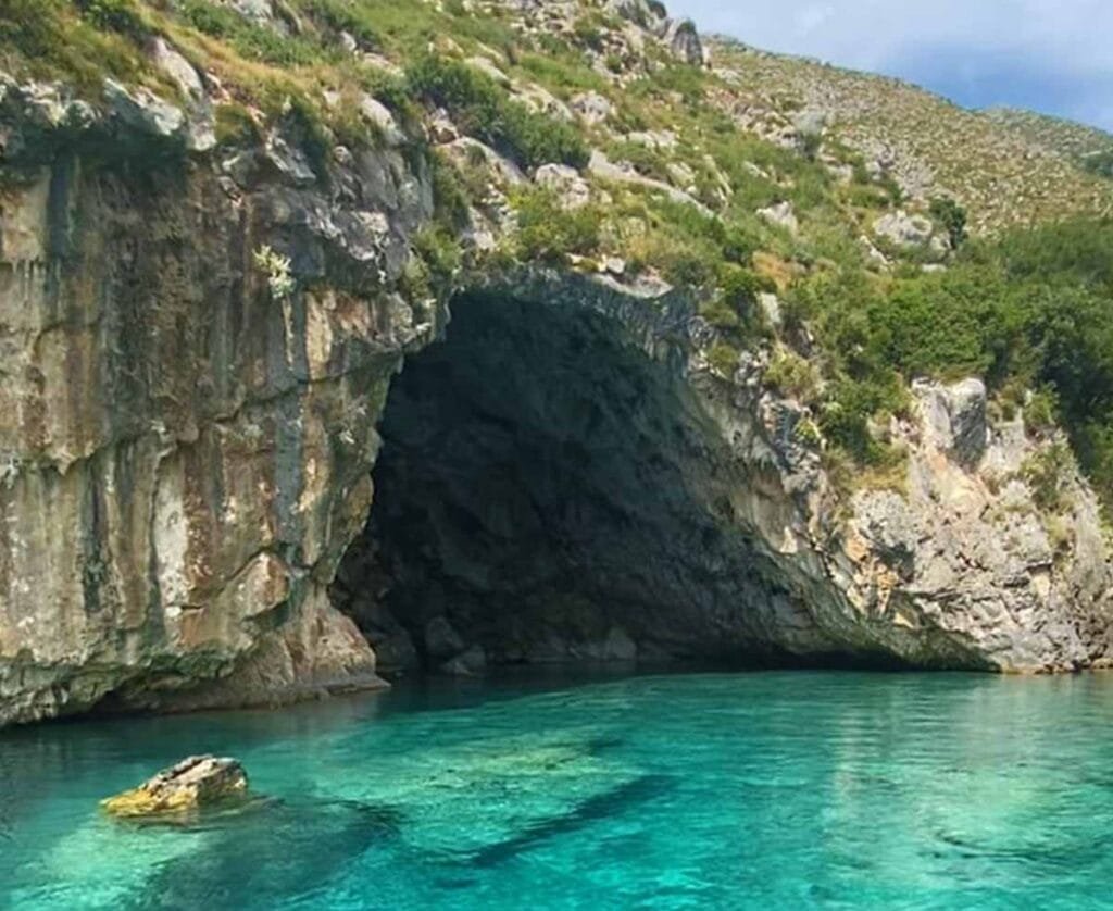 Church’s Cave in Vlore, Albania, with a wide rock entrance carved into limestone cliffs and crystal-clear turquoise water reflecting the cave’s dark interior along the Karaburun Peninsula coastline.