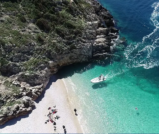 Aerial view of Dafina Bay with a white speedboat anchored in shallow turquoise water beside a small pebble beach, surrounded by rocky cliffs and clear emerald sea along the Karaburun Peninsula.