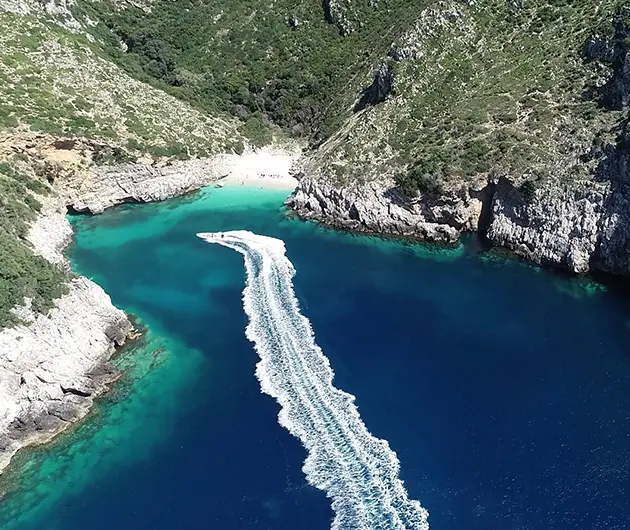Aerial view of Dafina Bay on the Karaburun Peninsula, showing a speedboat leaving a white wake across deep blue water, surrounded by steep rocky cliffs, green hillsides, and a small secluded beach with turquoise shallows.