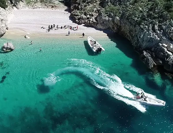 Aerial view of Dafina Bay on the Karaburun Peninsula, showing two speedboats in clear turquoise water near a secluded pebble beach, surrounded by rocky cliffs and lush green hills.