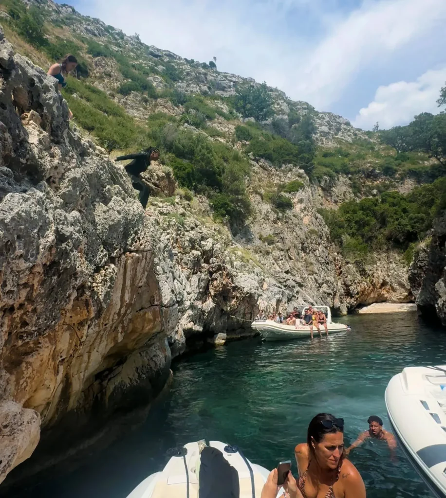 Visitors exploring Englishman Bay on the Karaburun Peninsula, with people jumping from rocky cliffs into clear emerald water while others relax on a speedboat anchored in the narrow rocky inlet.