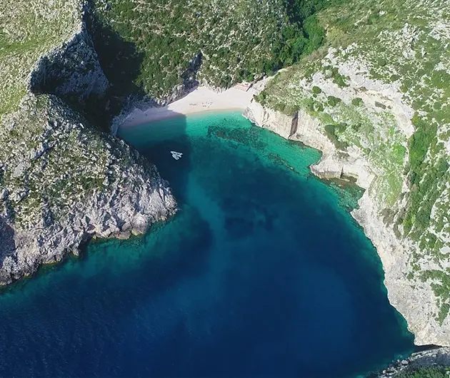 Aerial view of Grama Bay on the Karaburun Peninsula, showing a secluded pebble beach surrounded by steep white limestone cliffs and deep blue to turquoise water with a small boat near the shore.