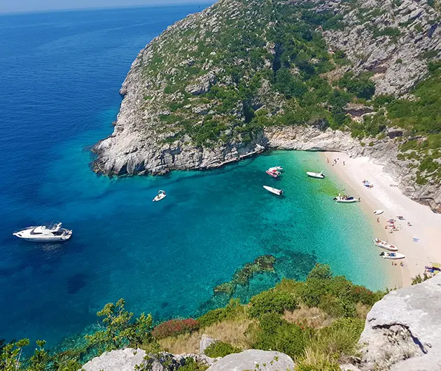Scenic view of Grama Bay on the Karaburun Peninsula, with turquoise water, several boats anchored near a white pebble beach, and steep rocky cliffs covered with green vegetation surrounding the bay.