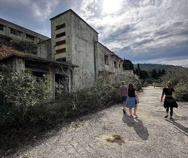 Visitors walking along a gravel road beside an abandoned military building on Sazan Island, Albania, with overgrown vegetation, concrete structures, and dramatic cloudy skies in the background.