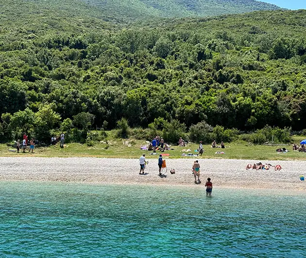 Shen Vasil Bay on the Karaburun Peninsula, featuring a quiet pebble beach with visitors relaxing and swimming, backed by dense green forest and rolling hills along the coastline.