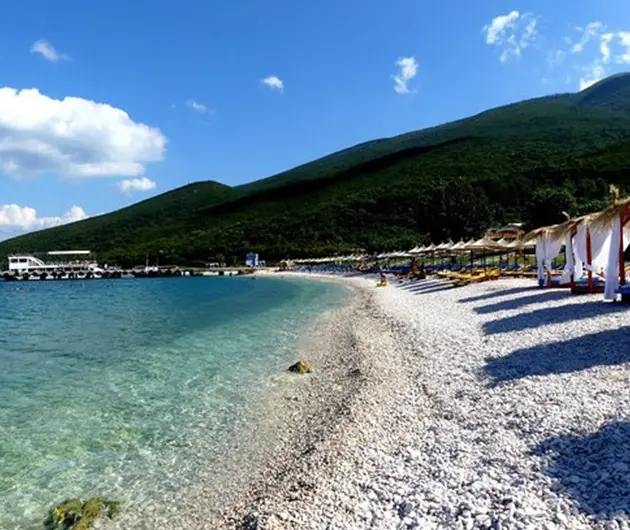 Shen Vasil Beach on the Karaburun Peninsula, with clear turquoise water along a white pebble shoreline, sun umbrellas arranged in a row, and green hills rising in the background under a bright blue sky.