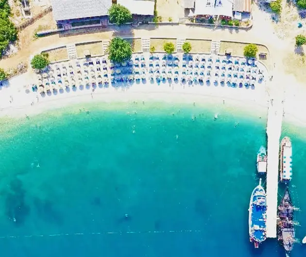 Aerial view of Shen Vasil Beach on the Karaburun Peninsula, showing a curved pebble shoreline lined with sun umbrellas and clear turquoise water, with boats docked at a small pier on the right side.
